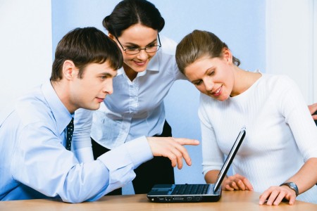 Portrait of serious businessman sitting at the table and pointing at the laptop monitor with two smiling ladies near by